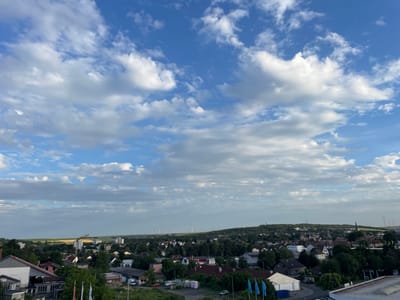 The image presents a panoramic view of Alzey, Germany, under a partly cloudy sky. The foreground shows the rooftops of buildings, including a large, light-colored structure with a sloped roof on the left, and other residential and commercial buildings with varying roof colors and architectural styles scattered throughout the mid-ground. Several trees are interspersed among the buildings, and some vehicles are visible on the streets and in parking areas.
In the mid-ground, a yellow construction crane stands tall near a cluster of buildings. Beyond this, a line of wind turbines extends across the horizon, indicating a rural or semi-rural landscape surrounding the town. The distant background features rolling hills covered in vegetation and a field of golden crops. The sky is a vibrant blue, populated by numerous white and grey cumulus clouds that cast shadows on the land below. The overall impression is one of a suburban or small-town setting on a pleasant, possibly late afternoon day.