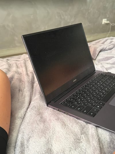 A laptop with a black screen and keyboard is open on a fluffy grey blanket. The laptop has the "HONOR" logo visible on its bezel. To the left of the laptop, a person's bare leg is visible, wearing black shorts. In the background, a grey wall has an electrical outlet with a white cord plugged into it. The scene appears to be indoors in a residential setting. No specific location within Novoaleksandrovka, Russia can be determined from the image.