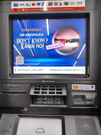 A dark gray Hyosung automated teller machine (ATM) is depicted in Bojongherang, Indonesia. The machine's screen displays an illuminated blue interface featuring a public service announcement. Prominent white text reads "#AWASMODUS LINK MENCURIGAKAN DON'T KNOW? KASIH NO!", with an image of a sunset over water circled by a red 'X' and a banner with "www.klikbc4.id" also marked by a red 'X'. A QR code for "Scan di sini" and the URL "bca.co.id/awasmodus" are visible. Two selectable options, "AKTIVASI KARTU ATM" and "TRANSAKSI TANPA KARTU," appear at the bottom of the screen. Above the screen, a red strip warns in Indonesian not to leave the machine prematurely, flanked by various bank and card network logos, including BCA, Visa, Plus, MasterCard, Maestro, JCB, GPN, ALTO, blu by BCA Digital, and BCA Syariah. Below the screen, the machine features a receipt dispenser slot, a cash dispenser with an integrated numeric keypad, a card insertion slot labeled "MASUKKAN KARTU ANDA," and a contactless payment symbol. No individuals are present in the image.