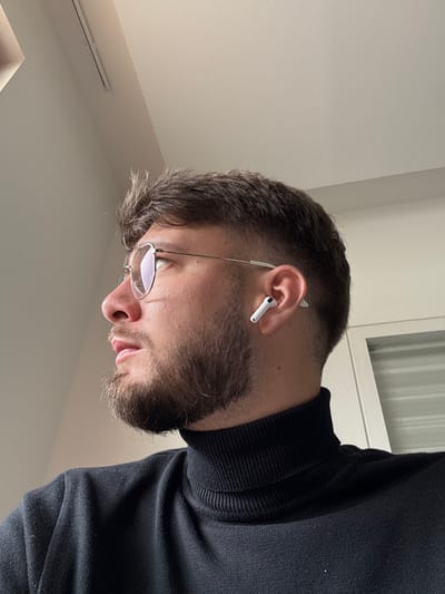 The image displays a male individual in a profile view, situated indoors in Lièpvre, France. The subject, a young adult with fair skin, dark short hair in a faded style, and a trimmed beard and mustache, is oriented to his left. He is wearing thin, round, metallic-framed eyeglasses with reflective lenses and a black ribbed turtleneck sweater. A white wireless earbud is inserted into his visible right ear. A minor scar or mark is present on the bridge of his nose, below the left eye. The immediate background consists of plain, light-colored walls and a ceiling. A linear vent or light fixture is partially visible on the ceiling in the upper left. A vertical white architectural element, consistent with a door frame or window frame, occupies the right edge of the frame.