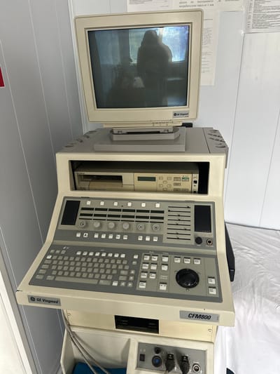 The image shows a vintage GE Vingmed CFM800 ultrasound machine. The machine consists of a CRT monitor, a central processing unit, and a control panel. The monitor is off, displaying a reflection of the surrounding environment. The CPU has a Sony color video printer and other internal components visible. The control panel is open, revealing a keyboard, sliders, knobs, and buttons. Cables are visible connecting parts of the system. The ultrasound machine is in a medical facility. A document with Bulgarian Cyrillic text is partially visible, referencing a "потребителска такса от 1 лев" (consumer tax of 1 lev) and text that contains a reference to "работоспособност" (work capacity) and "Zemen, Bulgaria". The surfaces surrounding the machine appear to be part of a medical room setting.
