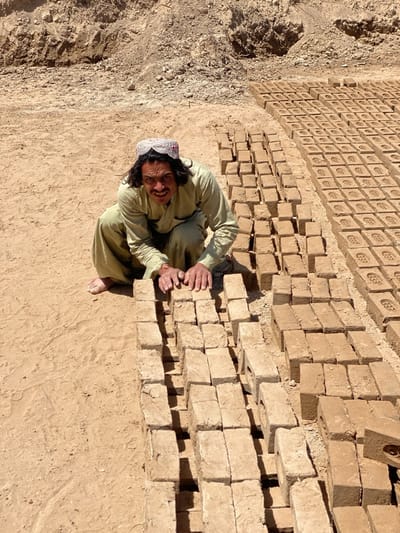 The image depicts a man crouching on the ground beside rows of newly made bricks in Deh-E Bagh, Afghanistan. He wears a traditional light-colored outfit and a decorative headpiece. His hands rest on a row of bricks in front of him. The bricks are uniform in shape and appear to be drying in the sun. The ground is arid and sandy, and a pile of earth is visible in the background.