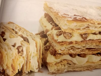 The image shows a close-up of a layered pastry, likely a mille-feuille or Napoleon, on a white surface. The pastry has multiple flaky layers of golden-brown puff pastry interleaved with a thick, pale yellow cream filling. Small, dark brown pieces, possibly nuts or chocolate chips, are visible within the cream layers. The top layer of the pastry is dusted with a fine white powder, which appears to be powdered sugar. The pastry is cut, revealing the distinct layers and the creamy filling. The background is a plain white, suggesting a neutral setting for showcasing the dessert. There are no people or any indication of a city, street, or any specific location within Rumaithiya, Kuwait. The image is solely focused on the dessert.