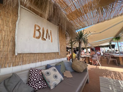 The image depicts an outdoor seating area of a beachfront establishment, likely a restaurant or bar, in San Roque, Spain. The focal point in the foreground is a brown upholstered bench with several cushions. One cushion features a geometric diamond pattern in white and dark brown, while another displays a black and white floral design on a light background. A large woven straw bag rests on the bench to the right. Behind the bench, a wall is constructed from vertical bamboo reeds, topped with a thatched roof made of dried palm fronds. A white sign with cut-out letters spelling "BLM" in a rustic, wooden style is mounted on the wall.
The background reveals more seating areas with wooden tables and chairs occupied by patrons. The overall ambiance is casual and breezy, with natural light filtering through the bamboo and thatched roof. Palm trees are visible, adding to the tropical, coastal feel. The ground appears to be sandy or dirt. The establishment seems to be open for business, with people dining and socializing, suggesting a daytime scene during pleasant weather. The lighting suggests it is daytime.