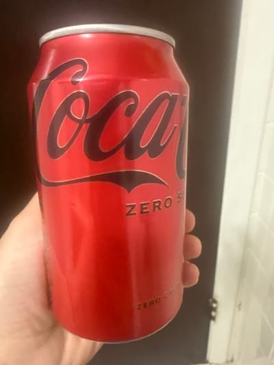 The image displays a close-up, horizontal view of a red Coca-Cola Zero Sugar can held by a hand. The iconic "Coca-Cola" script logo, rendered in black with a white outline, is prominently featured on the can's surface. To the right of the main logo, the text "ZERO SUGAR" is visible, stacked vertically in white lettering. Below "ZERO SUGAR," smaller white text clarifies "ZERO CALORIE."
A hand with fair skin tone is visible on the right side of the image, grasping the can from underneath. The fingers are gently curled around the can's curvature.
The setting appears to be indoors, with a dark, uniform surface, likely a table or countertop, occupying the mid-ground of the background. Above this dark surface, a lighter, off-white area suggests a wall or cabinet, with a subtle rectangular or tile-like texture visible. In the upper right corner of the background, faint metallic fixtures or hinges can be discerned, consistent with cabinet hardware.
The activity depicted is simply the holding of the beverage can. The lighting is even, suggesting an indoor environment without specific time-of-day indicators. No particular emotions are conveyed. The location context of Des Plaines, United States, is provided, but there are no specific visual cues within the image itself to confirm this geographical detail.