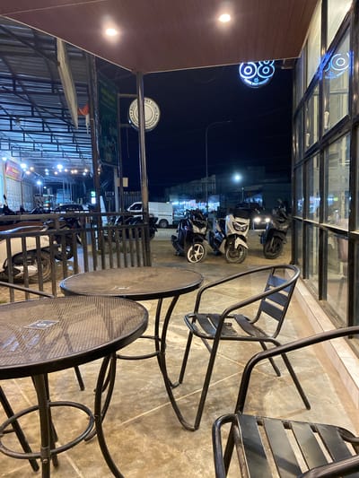 The photograph, taken at night, shows the exterior of a commercial establishment in Keudeublang, Indonesia. The scene is viewed from under an awning, with tables and chairs in the foreground. The furniture consists of metal-framed tables and chairs with a mesh design.
Outside the establishment, a row of motorcycles is parked along the curb. Across the street, lights from other businesses illuminate the area, including signs in Indonesian script. The reflections of these lights are visible in the glass windows of the building.
A neon sign for a "COFFEE SHOP PREMIUM" is visible on the building.