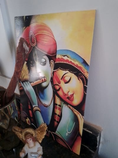 An indoor scene in the city of Campo Grande, Brazil, features a rectangular canvas print leaning against a light-colored wall. The print depicts the Hindu deities Krishna and Radha. Krishna, on the left, has blue-green skin, a red turban with beaded ornamentation, and a 'U'-shaped tilak on his forehead. He is shown playing a wooden flute with his hands. Radha, on the right, has fair skin, a blue head covering, and a red bindi on her forehead, with her eyes closed, leaning her head on Krishna's shoulder.

In the bottom-left foreground, a metallic faceted container holds a dark, textured, irregular object that extends upwards, partially obscuring the top-left portion of the print. To the right of the container, a small figurine of an angel with golden wings, fair skin, and reddish-brown hair, wearing a white robe and holding a small silver object, rests on a dark, reflective surface. The wall to the right of the print is plain and off-white. The bottom-right foreground reveals a dark, chipped, and dusty ledge or surface.