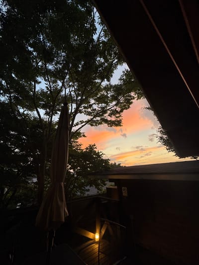 The image displays an outdoor scene at twilight from an elevated perspective in the Rokko Mountain area of Kobe, Japan, overlooking what appears to be Osaka Bay. The sky, occupying the upper two-thirds of the frame, exhibits a gradient of colors from deep orange and pink hues along the horizon to lighter blues and whites directly above, with scattered cloud formations. A large, leafy tree is silhouetted prominently on the left and center, its dense foliage obscuring parts of the sky. In the lower-mid left, a folded, light-colored outdoor patio umbrella stands vertically, also silhouetted. The foreground and right side of the image are framed by dark, architectural elements, consistent with a wooden structure or roof overhang. Below this, in the midground, a dark brown building with a sloped, dark-tiled roof is visible. A single, glowing yellow outdoor lamp is affixed to a wooden railing or post in the lower-mid right, providing localized illumination. In the distant background, a dark, expansive horizontal line is visible, indicative of a large body of water, consistent with Osaka Bay or the Seto Inland Sea. No individuals are present.