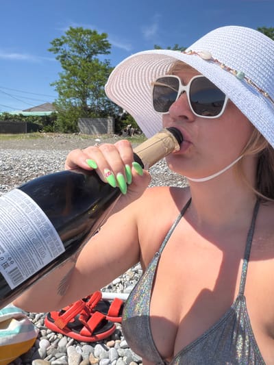 A woman is holding a bottle of champagne and appears to be drinking from it. She is wearing a white sun hat, sunglasses, and a sparkling silver bikini. Her fingernails are painted a bright green. The background shows a rocky beach with some trees and buildings. The sky is blue and clear, suggesting a sunny day. The scene appears to be a leisurely outdoor activity, possibly a celebration or a vacation. Text on the champagne bottle indicates it is from the Republic of Abkhazia, with specific details about the grape varieties, producer, and harvest year. The text also includes warnings about excessive alcohol consumption.