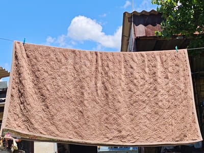 A brown towel with a textured pattern hangs on a clothesline outdoors. Two blue clothespins secure the towel to the line. The background consists of a blue sky with white clouds, a corrugated metal roof, some foliage from a tree, and parts of buildings. The scene appears to be in a residential area, likely a backyard, in Ароматненское сельское поселение, Ukraine.