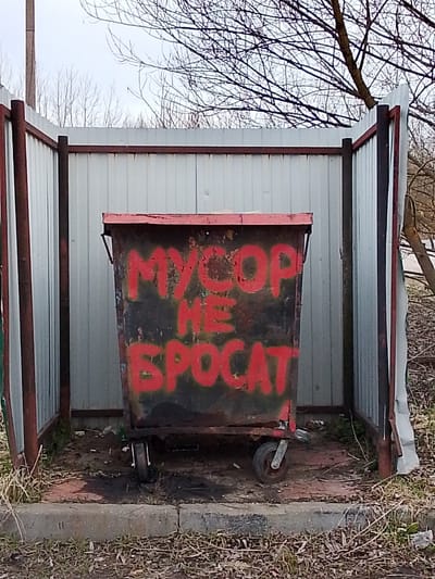 A medium, centered shot reveals a trash container within a fenced enclosure located in the city of Снопок Старый, Russia. The trash container, predominantly black, has the phrase "MYCOP HE SPOCAT" spray-painted in red. The container is equipped with wheels and appears to be in a state of disrepair. The enclosure surrounding the trash container features corrugated metal sheets and dark metal posts, showing signs of rust and weathering. The ground inside the enclosure is littered with debris. Outside the enclosure, the environment includes dry grass, bare branches, and a concrete curb. The sky above is overcast.