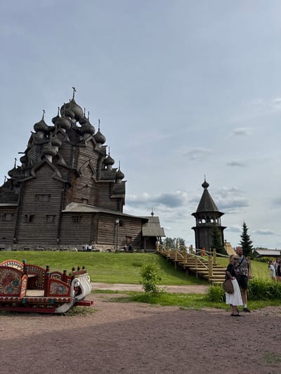 In Nevskii Parkleskhoz, Russia, a wooden church with multiple domes stands prominently against a cloudy sky. To the right of the church, a wooden bell tower with a conical roof is visible. A wooden staircase with railings leads up a grassy incline towards the church. In the foreground, a decorative, painted wooden sleigh is situated on a gravel path. Two individuals, a man and a woman, stand near the staircase, facing towards the church. Several other people are scattered in the background, some sitting on a bench in front of the church. The environment appears to be a park or outdoor historical site, with grass and trees surrounding the structures.