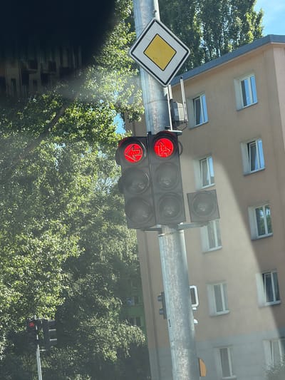 This media file captures a street scene in Olomouc, Czechia, likely during daytime. The foreground is dominated by a traffic light showing a red left-pointing arrow and a red downward-pointing arrow, indicating a mandatory left turn and straight ahead respectively. A yellow diamond-shaped traffic sign with a thick black border, signifying a "priority road" in many European countries, is visible to the left of the traffic light.

Above the traffic light, the facade of a building with several skylight windows is visible. The building appears to have a neutral-colored exterior, possibly beige or light gray, with a blue trim along the edge. Sunlight illuminates the scene, casting shadows and creating a bright atmosphere, suggesting fair weather. Lush green trees with dense foliage fill the background, obscuring other parts of the urban environment. A light metal pole or beam supports the traffic light and the sign, indicating typical road infrastructure. The image seems to be taken from a moving vehicle, given the slight blur and the angle of view, looking upwards. There are no people or animals clearly visible in the frame, focusing solely on the traffic control devices, the building, and the surrounding greenery.