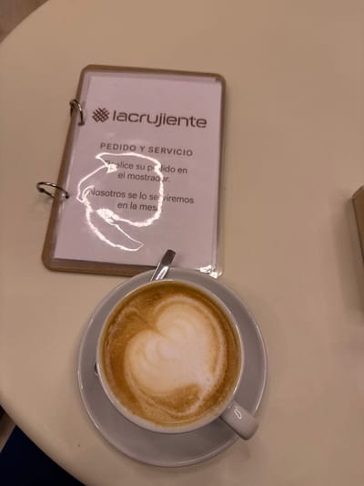 The image displays an overhead view of a table within a cafe or bakery identified as "lacrujiente" in Alcobendas, Spain. The primary subjects are a white ceramic coffee cup containing a latte with a distinct heart-shaped foam art, resting on a matching saucer with a silver spoon, and a small, rectangular sign holder. The sign holder features a light-colored frame, metal ring binders, and displays instructions in Spanish on a white sheet of paper protected by a clear plastic cover. The text reads: "lacrujiente" (logo), "PEDIDO Y SERVICIO" (ORDER AND SERVICE), "Realice su pedido en el mostrador." (Place your order at the counter.), and "Nosotros se lo serviremos en la mesa." (We will serve it to you at the table.). Bright, irregular reflections from an overhead light source are visible on the plastic covering of the sign. The table surface is a uniform light beige or cream color. A small, light brown cardboard object is partially visible on the right edge of the frame, and a dark fabric, possibly clothing, is visible in the bottom left corner. No individuals are depicted.