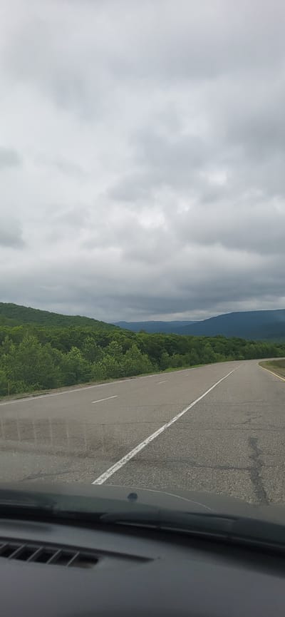 The image shows a road in Zanadvorovka, Russia, with lush green hills and mountains in the background. The sky is overcast with dark, heavy clouds, suggesting a stormy or rainy day. The road is asphalt and has white lines marking the lanes. The foreground shows the dashboard of a car, indicating the photo was taken from a moving vehicle. No people or other subjects are visible in the image. The environment is natural and appears to be a rural or mountainous area.