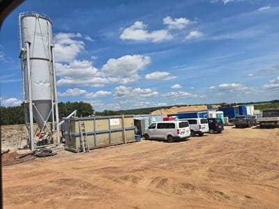 This image depicts an outdoor scene at what appears to be a construction or industrial site in Piecnik, Poland. The central focus is a large, grey cylindrical silo, likely for storing materials, which is situated on the left side of the frame. Adjacent to the silo is a long, metallic rectangular container with a ladder attached, suggesting it is used for storage or as part of a processing unit. Several vehicles are parked in the background, including white vans, a dark-colored SUV, and a pickup truck. Interspersed among the vehicles are shipping containers, some blue and some white. Piles of material, possibly wood or sand, are visible further back, along with a line of trees forming the distant horizon. The ground is composed of reddish-brown dirt with tire tracks, indicating recent vehicle activity.
The sky is bright blue with scattered white cumulus clouds, suggesting fair weather and daytime, likely mid-morning to afternoon. The sunlight casts shadows, further indicating the time of day. There is no clear activity or event taking place; the scene appears to be a static view of the site.
Notable details include the weathered appearance of the silo and the storage container, hinting at the functional nature of the location. The presence of various containers and vehicles suggests logistical operations. Visible text is limited; a license plate is partially visible on one of the vans, and a logo or small label is on the back of another van. No people are clearly visible in the image. The overall impression is one of an active, albeit currently quiet, industrial or construction yard under a pleasant sky.