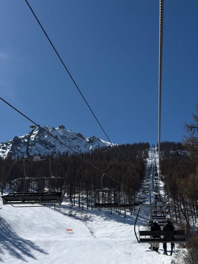 The image captures a winter mountain scene from the perspective of a ski lift ascending a slope in Saint-Chaffrey, France. In the immediate foreground, two parallel steel cables extend diagonally across a clear, deep blue sky.

On the right side of the frame, a chairlift chair, identified by the number "110" on its frame, transports two individuals uphill. Both wear winter ski attire; the left individual has a dark jacket, and the right wears an olive-green jacket. Their skis are visible hanging below the chair, indicating they are equipped for skiing. They face away from the camera, towards the upper part of the slope.

To the left of the occupied chair, another empty chairlift chair, numbered "138," is visible, hanging over a wide, groomed ski slope covered in white snow with visible ski tracks. A small orange marker is present on the slope's surface.

The mid-ground features a dense forest of dark-trunked, bare-branched trees covering the mountainside. Further up the slope, a continuous line of chairlift chairs and their supporting pylons ascend the mountain, extending into the distance along the treeline. In the background, snow-capped rocky mountain peaks rise under the bright, clear sky. Strong sunlight casts distinct shadows across the snowy terrain.