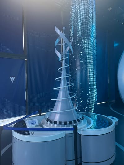 An indoor exhibition space in Guangming District, China, is illuminated primarily by blue light. The central object is a silver-colored, futuristic display model, comprising a conical base, encircled by an upward-spiraling ramp, and topped with two curved, blade-like forms and a sphere. This model is mounted on a multi-tiered circular base in white and grey. On the left side of the base, a white informational sign with Chinese text, "中科创新智" (Zhongke Innovation Intelligence), is displayed. To the right, a semi-circular basin integrated into the base contains numerous small blue spheres submerged in a clear liquid. A black stanchion with a blue retractable belt forms a barrier on the left side of the exhibit. The background consists of dark blue wall panels; one on the left displays a white triangular sign with a fire hydrant icon and Chinese characters "消防栓口" (Fire Hydrant). Behind the main model, a tall, vertical column of bright blue LED dots generates a dynamic, sparkling effect. A partial view of a large, light-colored, curved object is visible on the far right. No individuals are present in the scene.