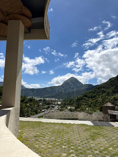 The image presents a view in Sembalun Bumbung, Indonesia, featuring a mountainous landscape under a partly cloudy sky. A settlement nestled at the base of the mountain is visible. In the foreground, architectural elements such as pillars and parts of a building structure are present. Adjacent to the building, there's a section of grass and concrete blocks.