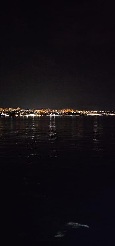 The image depicts a nighttime view of the coastline of Split, Croatia. A city skyline, illuminated by numerous lights, stretches across the horizon. The lights are a mix of warm yellow and white, indicating buildings and streetlights. The city is situated on a landmass that meets the dark, rippling water in the foreground. The water reflects the city lights, creating shimmering streaks across its surface. The sky above is dark and largely featureless, suggesting it is either night or heavily overcast. The lower portion of the image is dominated by the dark water, with a faint, pale form visible near the bottom right, possibly a reflection or a submerged object. No people or specific activities are discernible. The scene conveys a sense of urban tranquility at night, viewed from across the water.
