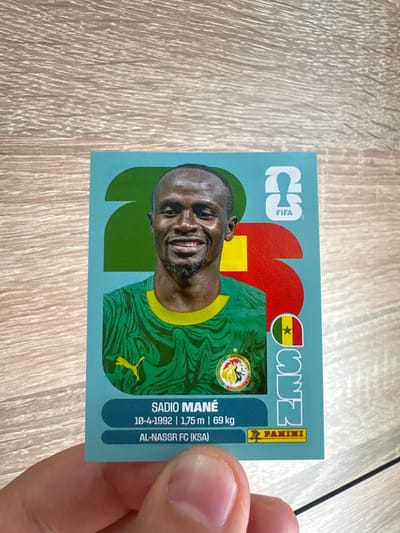 A man is holding a collectible card featuring a portrait of soccer player Sadio Mané. The card is light blue with large graphic elements in red and green, as well as the FIFA logo. Mané is wearing a green soccer jersey with a yellow trim and a yellow Puma logo. His personal information, including his name, date of birth, height, weight, and club (Al-Nassr FC), is printed on the card, along with the Panini logo. The background of the image is a light brown wooden surface. The man's hand is partially visible at the bottom of the frame, holding the card.