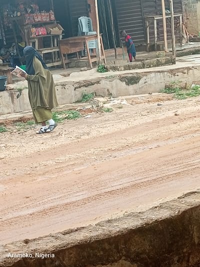 The image shows a dirt road in Atamoko, Nigeria (latitude: 7.834, longitude: 4.479). A woman wearing a long green robe and a head covering is walking down the road, holding a book. She is walking towards the right side of the image. There is a small child in a red shirt and blue shorts, visible in the background, leaning over and looking down at something on the ground. The road is unpaved and dusty. The ground is covered in dirt, and the edges of the road are lined with concrete. There are some buildings and shops in the background, as well as some vegetation. The scene appears to be in a residential area.