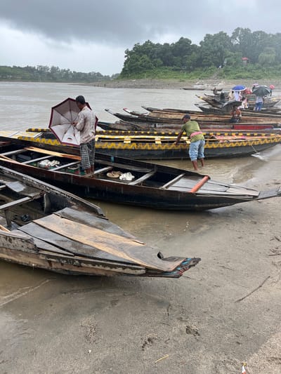 The image depicts a riverside scene in Jaflong, Bangladesh, with several wooden boats docked along a sandy bank. The sky is overcast, suggesting rain, and a light drizzle appears to be falling, as indicated by water droplets on the boats and the presence of umbrellas.

In the foreground, a man in checkered pants and a light-colored shirt stands holding a partially opened umbrella. He is standing in the water near a wooden boat, with several pairs of sandals visible inside it. To his left, another wooden boat is partially submerged and appears damaged.

Further back, more boats are lined up along the shore. Some of these boats are occupied by people, many of whom are holding umbrellas of various colors, including blue and black. One man in a green shirt and blue pants is visible near the water's edge, tending to one of the boats.

The background features a lush green hillside covered in trees, with a few small buildings visible. The river is wide and murky, with a gentle flow. The overall atmosphere is one of a functional, albeit somewhat weathered, riverine transportation hub operating in rainy weather.