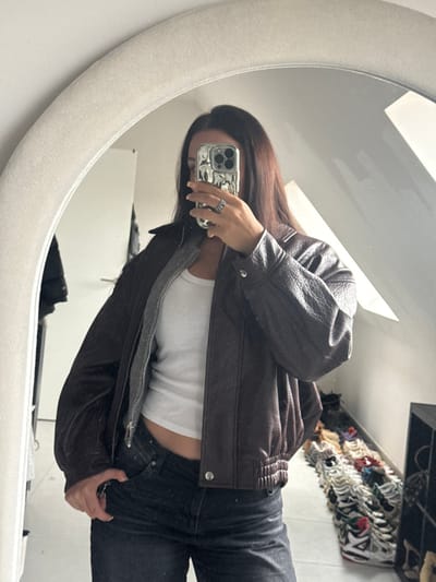 A subject, identified as female, with long dark hair, is depicted in a mirror selfie taken within a room in Tielrode, Belgium. The individual holds a smartphone with a distinctive chrome-like case in their right hand, obscuring their face, while a ring is visible on the right ring finger. The subject wears a dark brown, textured bomber-style jacket unzipped over a white ribbed tank top and dark-wash jeans. The environment, visible in the mirror's reflection, includes a room with a light-colored floor and walls, a sloped ceiling indicative of an attic space, and a rectangular window or skylight admitting natural light. To the right of the subject, a substantial collection of various sneakers is arranged in multiple rows on the floor, adjacent to a dark, cubby-style shelving unit. To the left, a portion of a light-colored wardrobe or door is visible, with a dark garment hanging. The mirror itself features a thick, light-colored, textured arched frame.
