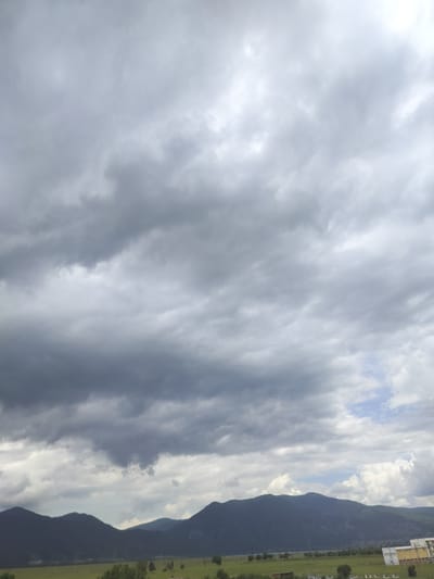 The image depicts a view of the landscape and sky near Gorno Sahrane, Bulgaria. In the foreground, a green field stretches towards a range of dark, silhouetted mountains. A few scattered trees are visible in the field. In the distance, a collection of buildings, possibly a town or industrial area, can be seen nestled against the foothills of the mountains. The sky is dominated by thick, gray, and white clouds, suggesting an overcast or potentially stormy day. Patches of lighter blue sky are visible in some areas between the clouds. The overall scene is one of a natural landscape under a dramatic sky.