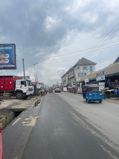 This image captures a bustling street scene in Rumuokwuta, Nigeria, under an overcast, cloudy sky. The perspective is from the roadside, looking down a paved road that stretches into the distance, flanked by commercial buildings.

In the foreground on the left, a large billboard advertises "ANGEL MISSION" with information for "BIBLE STUDY" (Wednesday, 6:00 PM) and a "HEALTH CLINIC" (Tuesday, 10:00 AM), specifying "Mgbouba Port Harcourt" and "www.alemembassy.org." Below the billboard, a white truck with a large red container filled with red crates is parked or moving slowly alongside the road.

The asphalt road, showing signs of wear and damp patches, occupies the center and right side of the frame. A blue three-wheeled auto-rickshaw, branded "TVS," is prominent on the right side, driving away from the viewer. Further down the road, other vehicles, including cars and more auto-rickshaws, are visible, indicating ongoing traffic.

Buildings line both sides of the street, many featuring awnings and signs for various businesses. One notable building has a distinctive roof and several blue-tinted windows. Visible text on shop signs includes "CJRE URCES," "TESSYGOLR," and "POS," as well as "ELECTRONIC, ELECTRICAL & GENERAL MERCHANT." A complex network of utility and power lines crisscrosses the sky above the street. The scene depicts a typical daytime urban environment with commercial activity and vehicular movement.