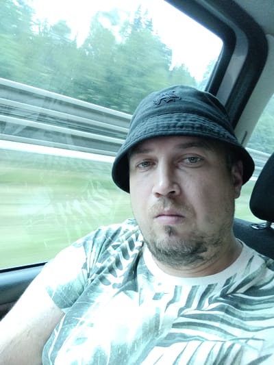 A man wearing a black bucket hat and a t-shirt with a leafy pattern is pictured from the chest up, with his face looking towards the camera. He has light skin, short brown hair, and a light beard. He appears to be in a vehicle, as indicated by the window and the interior of a car or train visible in the background. The window shows a blurry green landscape with trees, suggesting the vehicle is in motion. The lighting is bright, but the sky appears overcast, implying a cloudy day. The overall mood is casual and relaxed. There is no visible text in the image. The location context provided is Ostředek, Czechia.