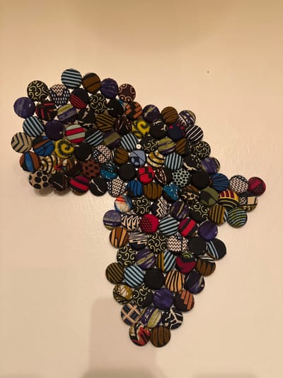 A colorful arrangement of round fabric buttons forms an abstract image resembling a horse's head. The buttons, in varying sizes and patterns, are closely packed together against a light tan background. The buttons feature diverse patterns including stripes, polka dots, and geometric shapes in shades of red, blue, black, and brown. Some buttons are flat, while others are slightly domed. A single shiny object is present in the lower portion of the arrangement.