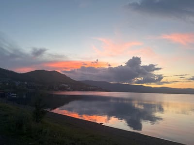 This is a landscape photograph of a tranquil lake at sunset, likely located in the vicinity of the city of Якты-Куль, Russia. The sky is a canvas of soft blues and pinks, with scattered clouds catching the last rays of the setting sun, casting a warm glow on the horizon. In the distance, rolling hills covered in lush vegetation and dotted with a small settlement of houses line the opposite shore of the lake. The water's surface is exceptionally still, acting as a mirror reflecting the vibrant colors of the sky and the silhouetted forms of the distant hills and clouds. The foreground features a gently sloping shore made of pebbles and grass, with a few sparse plants. A small, dark tree stands in the mid-foreground on the left side of the frame, adding a touch of depth to the scene. A few boats are moored along the shoreline on the far left. The overall atmosphere is one of peace and serenity.