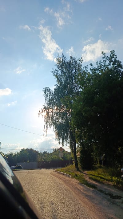 This media file depicts a bright, sunny day in a rural or suburban area, likely Zvenigorod, Russia. The scene is dominated by a clear blue sky with scattered white clouds, and the sun is shining brightly, casting a warm glow and creating lens flare. The sun's rays are partially obscured by the leaves of a large tree in the foreground, a birch tree with its characteristic thin trunk and drooping branches, and a dense, dark green tree next to it.
The foreground features a gravel road that curves to the right, suggesting a turn or intersection. To the left of the road, a grey fence separates the road from what appears to be a residential area with houses and more trees visible in the distance. A power line runs horizontally across the mid-ground of the image. To the right of the road, there is a patch of green grass and undergrowth, with the large trees casting dappled shadows.
A road sign with a yellow diamond shape and a white arrow pointing to the right is visible near the trees on the right side of the road. Below the arrow, there is some text in Cyrillic script, which appears to read "ул. Парковая" (ul. Parkovaya), indicating "Park Street."
The overall impression is one of a peaceful, warm summer day in a village or a quiet neighborhood on the outskirts of a town. The bright sunlight and clear sky evoke a sense of warmth and tranquility. There are no people visible in the media file, but the presence of a road and houses suggests human habitation. The image seems to have been taken from a moving vehicle, given the perspective and the blurry dark object in the lower left corner, possibly part of a car.