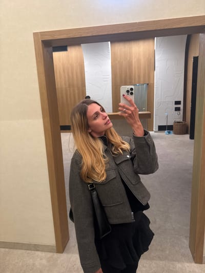 A blonde woman with long, wavy hair is taking a mirror selfie. She is wearing a grey, cropped jacket with a zipper and two chest pockets, over a black top and a black ruffled skirt. She is holding a white smartphone in her right hand and looking up and to the right. A black shoulder bag is slung over her left shoulder. The background appears to be a hallway or room with light-colored walls and a wooden doorframe. The flooring is tiled. The reflection in the mirror shows a clean, modern space with wood-paneled walls and some abstract white wall art. There are also some cleaning supplies visible in the reflection, including a dustpan and broom. The scene is well-lit and appears to be indoors.