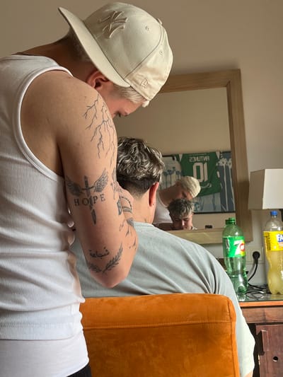 A close-up shot depicts two individuals partially visible through a mirror's reflection. The person in the foreground wears a white tank top and a light-colored baseball cap with the New York Yankees logo. Their left arm is adorned with several tattoos, including lightning bolts and a sword with the word "HOPE." The second person visible through the mirror also wears a similar cap and tank top. Green and yellow bottles are on a shelf visible above the mirror's reflection, near an orange piece of furniture. The mirror has a wooden frame and is set against a beige wall.
There is no information in the image to identify the location as Punta Umbría, Spain.