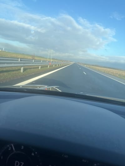 The image depicts a perspective from inside a vehicle traveling on a multi-lane highway near Воденичане, Bulgaria, under a partly cloudy sky. The foreground shows the top of a dark dashboard and the lower edge of a windshield, with reflections visible on the glass. A portion of a circular control dial displaying "D7" is visible on the lower left of the dashboard.
The midground features a straight, dark asphalt highway with at least two visible lanes extending into the distant horizon. The road surface appears wet or reflective. White solid lines mark the outer edge of the highway, and white dashed lines delineate the lanes. To the left of the highway, a metal guardrail system with silver rails and grey posts runs parallel to the road. Orange reflectors are spaced along the top of these guardrail posts. A grassy embankment rises beyond the guardrail.
The background reveals an open, flat landscape of green and brown fields under a wide blue sky dotted with scattered white and light grey clouds. A tall, slender structure, possibly an industrial tower or wind turbine, is faintly visible on the far left horizon. No individuals are present in the visible scene.
