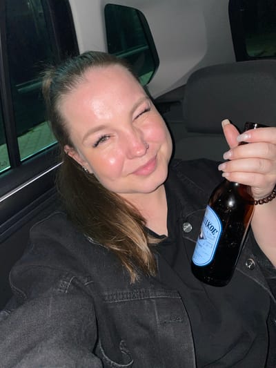 The image shows a woman holding a bottle of beer, smiling at the camera. She is wearing a black denim jacket. The beer bottle has a blue label with Russian text. The woman has a nose ring and her nails are painted with a shimmery polish. Her hair is brown with some lighter streaks. The background appears to be inside a vehicle, possibly a car, with a window visible. The lighting suggests it is nighttime or indoors. The scene conveys a sense of casual enjoyment.