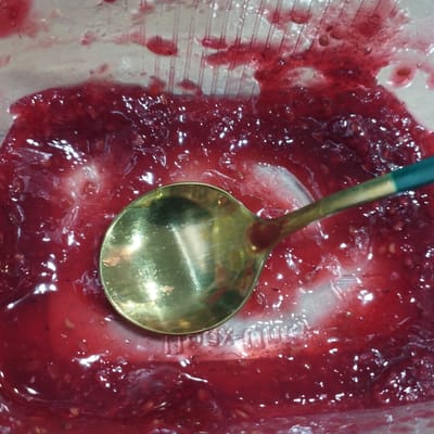 A close-up, overhead shot shows a golden spoon partially submerged in a viscous, translucent red substance, possibly jam or compote. The substance contains small seeds and fruit particles, with visible air bubbles and some lighter, cloudier areas. The spoon has a dark green handle. Underneath the substance, the ribbed texture of a clear plastic container is visible. The scene is domestic, likely a kitchen, and there is no indication of location in Покосное, Russia.