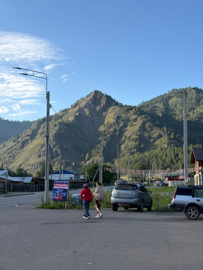 This is a vertical, eye-level, outdoor, full-shot photograph taken in the city of Uznezya, Russia. The scene depicts a wide, paved road with a slight upward slope in the foreground, leading towards a backdrop of lush green mountains. Two individuals are walking across the road from left to right. The person on the left wears a red jacket and blue jeans, while the person on the right wears a beige jacket and dark shorts. A light blue SUV is parked on the right side of the road, facing away from the viewer, and a silver SUV is parked to its right, also facing away. A tall, concrete lamppost stands to the left of the individuals, with power lines extending from it. A sign is posted on a lamppost to the right of the pedestrians, featuring an advertisement for river rafting excursions, with a phone number and Russian text. In the background, there are residential buildings with wooden fences and a dense forest covering the mountainside. The sky is a clear, bright blue with a few scattered white clouds. The overall lighting suggests it is either early morning or late afternoon, with a warm, natural light illuminating the scene.