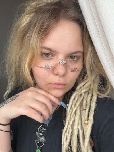 A young woman with blonde hair and dreadlocks, wearing a black t-shirt and a silver nose chain, is shown in a close-up portrait. She has a tattoo on her left arm and is holding her chin with her right hand. The background is a neutral gray with a white curtain on the right side. The location is not specified in the image.
