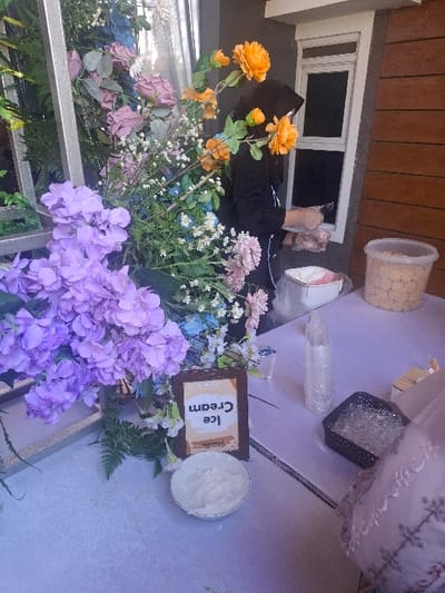 The scene takes place at a stall near the coordinates -6.968, 107.651 in the city of Bojongsoang, Indonesia. There is a woman wearing a black hijab standing behind a white table with a variety of objects on it. There is a bowl of white cream labeled "Ice Cream" next to a clear plastic bottle. Behind the table, there is a decorative arrangement of purple, white, and yellow flowers. The woman is holding a white container filled with something. Next to the woman, there is a container of snacks, an unidentified white container, and a black container filled with clear plastic wrapping. The wall behind the table appears to be made of concrete and wood. There is also a window with a black frame. The wall is adorned with a wooden plank structure. The environment is outdoors. The woman is standing near a white table, likely selling ice cream to customers.
