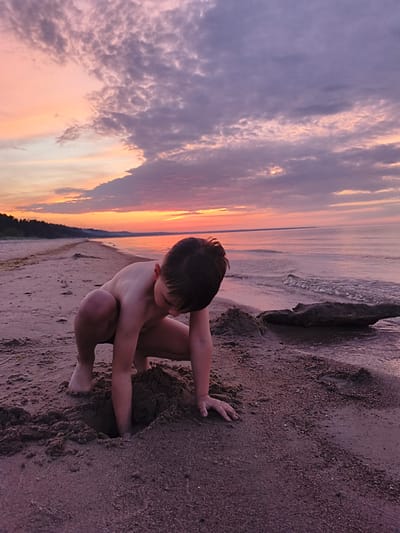 A young boy with dark hair is digging a hole in the wet sand on a beach at sunset in Apšuciems, Latvia. The sky is filled with pink and purple clouds. The ocean is calm and reflects the colors of the sky. The boy is wearing dark swim trunks. He is focused on digging. The sand is dark and damp, with small shells and pebbles. There is a piece of driftwood to the right of the boy. In the background, a line of trees can be seen along the coast. The overall impression is one of peaceful solitude and childhood innocence.