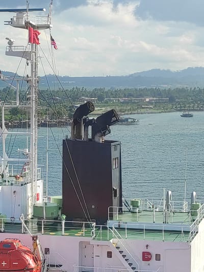 This media file captures a daytime coastal scene in Kunak, Malaysia, viewed from the deck of a large vessel, likely a cargo ship or tanker. The image is oriented vertically (sideways).

In the foreground and right side, the ship's superstructure is prominently featured, displaying white and green sections, railings, and a set of stairs. A large black exhaust stack with two pipes stands centrally. On the far right, an orange lifeboat with a white cross symbol is visible. To the left, a mast rises with rigging, antennas, and two flags – one red and another smaller pinkish-red flag. A red fire safety cabinet with indistinct markings can be seen on the superstructure.

Beyond the ship, calm, blue-green water extends towards a lush green coastline. Several smaller boats or pontoons are scattered across the water near the shore, which is densely covered with tropical vegetation. In the background, distant mountains or hills, also verdant, rise under a bright blue sky with scattered white clouds. The weather appears clear and sunny. No people are visible in the scene.