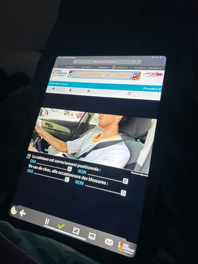 A digital tablet is displayed in a dimly lit environment, located in Arthès, France. The screen exhibits the `eleve.codesrousseau.fr` website, featuring a driving instruction multiple-choice question (number 10). The upper portion of the question presents an image of a male individual, appearing to be in his 20s or 30s with short dark hair, seated in the driver's seat of a car. He wears a white short-sleeved t-shirt with a yellow collar. His left arm extends to grip the steering wheel. A dark shoulder seatbelt is visible, incorrectly positioned across his neck rather than his shoulder. Through the car window, an outdoor scene with other vehicles and daylight is visible. The question text, in French, asks: "10 La ceinture est correctement positionnée :" (Is the belt correctly positioned :) and "En cas de choc, elle occasionnera des blessures :" (In case of a shock, will it cause injuries :). Response options A: OUI, B: NON, C: OUI, and D: NON are presented, with a note indicating "Plusiers réponses" (Multiple answers). The website header includes the "CODES ROUSSEAU" logo, a "CALENDRIER DE L'AVENT" banner, and the logged-in user's name, "GOGINASHVILI Salome." Browser tabs are visible above the main content, some displaying Georgian script. The tablet's status bar shows the time as 11:01, the date as Tuesday, December 30th, and a 19% battery charge. A prominent green checkmark icon is present within the website's bottom navigation bar. The tablet rests on a dark, possibly fabric, surface, from which screen reflections are apparent. The immediate ambient environment is characterized by minimal illumination.