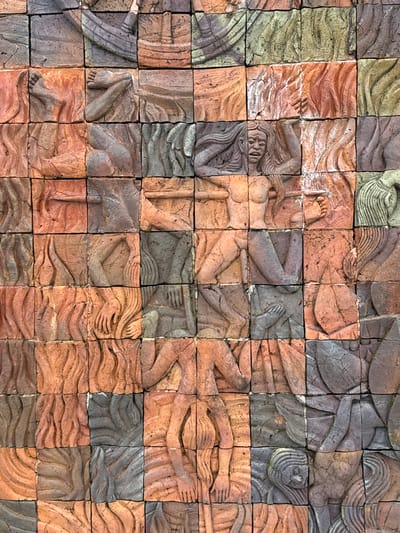 This image depicts a detailed, textured brick wall relief sculpture in Chang Khoeng, Thailand. The artwork is composed of numerous square bricks, each meticulously carved to form fragmented figurative elements and swirling patterns. The central focus is on human forms, with exposed torsos, limbs, and faces partially revealed within the textured surfaces. Many of the figures appear to be in dynamic or emotional poses, suggested by contorted limbs and strained expressions. The bricks themselves vary in color, ranging from terracotta oranges and reds to earthy browns and muted grays, contributing to the sculpture's aged and organic appearance. The rough, cracked surface of the bricks adds to the tactile quality of the artwork. The overall impression is of a fragmented narrative or a collection of interwoven human and abstract forms, presented in a communal outdoor setting.