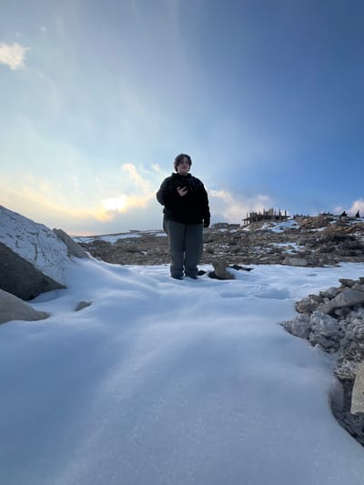 The image displays an outdoor, snow-covered, rocky landscape within the city of Talfita, Syria, under a partly cloudy sky. A single individual, appearing to be a young person or adult with dark, shoulder-length hair, stands centrally in the midground. This individual is dressed in a black hooded sweatshirt featuring a light speckled pattern on the chest, and dark grey trousers. A dark-colored object, consistent with a smartphone, is held in their right hand at chest height. The foreground is primarily covered by a textured layer of white snow, with large, light-colored rocks prominent on both the left and right edges. Beyond the individual, the terrain consists of sparse, rocky ground interspersed with smaller snow patches. In the distant background, to the right and on an elevated section, a multi-level concrete building structure under construction is visible. The sky transitions from a bright, partially obscured sun on the left to a clear blue on the right, populated by scattered white clouds.
