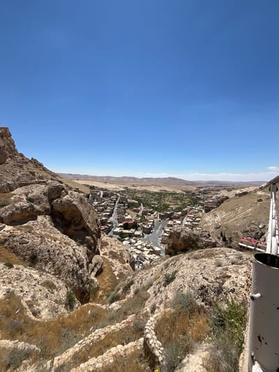 The image depicts an elevated view of Ma'loula, Syria, taken from a rocky, mountainous vantage point. A clear, blue sky occupies the upper portion of the frame. Below the sky, a line of arid, tan-colored land transitions into the urban sprawl of Ma'loula. The town consists of densely packed buildings, with visible roads running through the central area. The architecture primarily features earth-toned buildings with red roofs. The foreground is dominated by the rugged terrain of the mountain, consisting of light brown rock formations with sparse vegetation. In the upper right corner, a portion of a metallic structure is visible, likely part of the observation point's infrastructure.