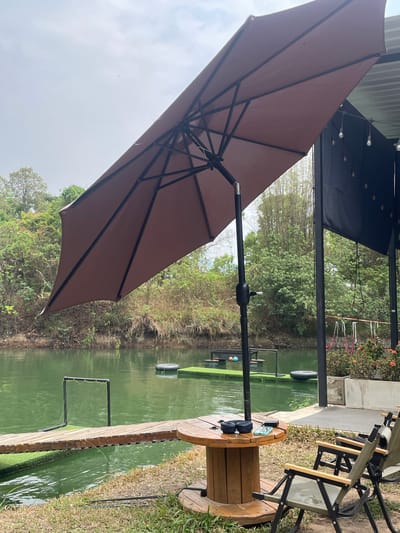 The image shows an outdoor scene at what appears to be a fishing or recreation area, potentially within Tambon Nam Phrae, Thailand. A brown umbrella is prominently featured on the left side of the image, offering shade. To the right, there is a wooden spool repurposed as a table, with items such as headphones and a phone on top. Next to the spool table, there are foldable chairs. Beyond the chairs and spool, there's a body of water with green tint, possibly a pond or lake. A wooden pathway or dock extends into the water. The landscape includes vegetation and trees in the background. Other floating objects such as tires and nets, or frame structures are in the water. The lighting suggests it's daytime, likely overcast.