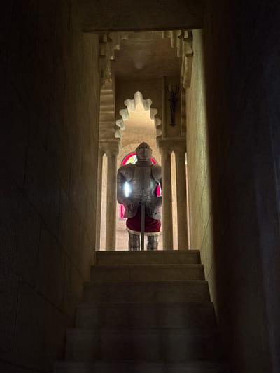 This image depicts an indoor scene, likely within a monument or historical building, given the stone architecture and the presence of a suit of armor. The setting is dimly lit, with strong shadows cast by the architecture, suggesting it is either nighttime or the interior is not well-illuminated. The primary subject is a full suit of medieval-style armor, positioned upright and seemingly on display. The armor is metallic, with visible articulation at the joints. It appears to be resting on a red cloth or cloak.

A notable feature is the reflection of the armor and its surroundings on a polished surface below, creating a mirrored image that is also visible in the frame. This reflection suggests the floor or a display base is highly reflective. In the background, behind the armor, there's a hint of a red stained-glass window, casting a colored glow. To the left of the armor, a dark, intricately shaped object, possibly a weapon or decorative artifact, is mounted on the wall. The architecture itself is characterized by stepped, carved stone elements, resembling stairs or decorative ledges. The overall mood is somber and historical, with the lighting and the subject matter evoking a sense of past grandeur or solemnity. The location context provided, Benalmádena, Spain, might suggest this is part of a local attraction or monument. No text is visible in the image.